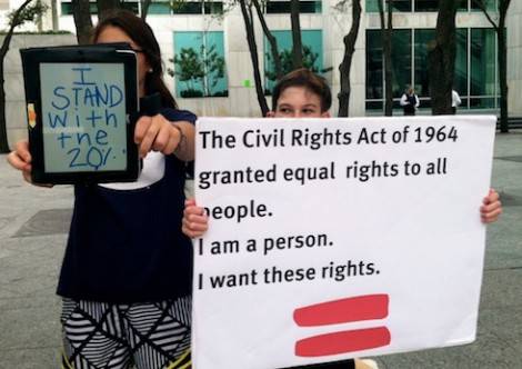 Girl with brown hair , blue tshirt is holding an iPad out in front of her. Her face is obscured by the iPad. Boy with Brown Hair holds a white sign that says " The Civil Rights Act of 1964 granted equal rights to all people. I am a person. I want these rights.