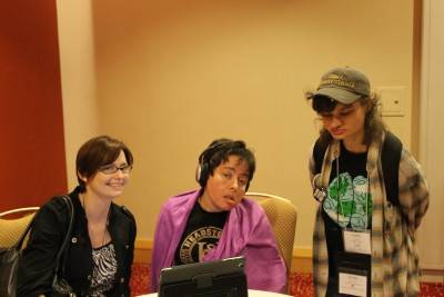 Photograph of three people in front of a table that has an iPad on it. The first person on the left is sitting down,has brown air, white skin, and is wearing glasses and a black sweater. The person in the middle is sitting down, has black hair, is wearing headphones and a pink sweater and black t-shirt. The third person is standing, has brown hair, is wearing a cap, and plaid shirt over a black t-shirt.