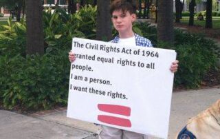 Boy with brown hair and blue and white checked shirt holds sign that reads The Civil Rights Act of 1964 granted equal rights to all people. I am a person. I want these rights.