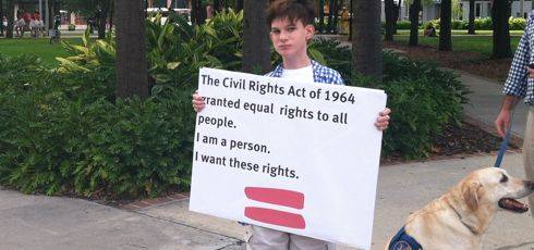 Boy with brown hair and blue and white checked shirt holds sign that reads The Civil Rights Act of 1964 granted equal rights to all people. I am a person. I want these rights.