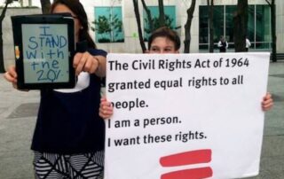 Girl with brown hair , blue tshirt is holding an iPad out in front of her. Her face is obscured by the iPad. Boy with Brown Hair holds a white sign that says " The Civil Rights Act of 1964 granted equal rights to all people. I am a person. I want these rights.