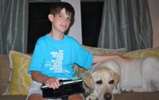 boy wearing a turquoise tshrt that reads "Not being able to speak is not the same as not having anything to say" sitting on couch one hand on iPad one hand on his dog . The dog is yellow lab/golden mix .