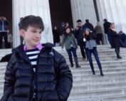 a teenage light skinned boy with freckles, wearing a black jacket, blue and white striped sweater, pink collared shirt is standing in front of large columns and steps that lead to the Lincoln Memorial