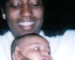 Photograph of woman with brown skin and dark brown shoulder length hair looking down smiling while holding her baby.