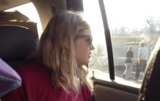 Photograph of Issy Stapleton. Light skinned blonde girl with shoulder length hair looks out a car window. She is wearing sunglasses and a pink tshirt.