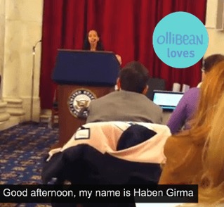 Image description: Photograph taken from back of a room . Picture shows audience members backs as they look toward person with long brown hair standing at a lecturn. There is a maroon curtain behind speaker. In upper right hand of photograph a turquoise circle with purple text that reads " ollibean loves". At bottom of photograph, closed captioning text in white against a vlick background reads " Good afternoon, my name is Haben Girma."