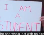 Image description. Hands holding up a white sign with pink text " I AM A STUDENT" Closed captioning text" I'm not the one who's lost with no direction, no"
