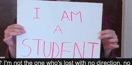 Image description. Hands holding up a white sign with pink text " I AM A STUDENT" Closed captioning text" I'm not the one who's lost with no direction, no"