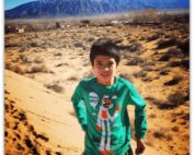 Image of young boy with olive skin and black hair smiling. He is standing in the desert.